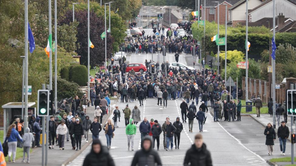 A wide angle shot showing large crowds on the Garvaghy Road, split out onto two distinct groups, spread across the length of the road, blocking access. There are also some vehicles parked across the road, again blocking it. The street lamps lining the road, which features greenery and red bricked homes, are adorned with Irish green-white-orange tricolor and blue starry plough flags.