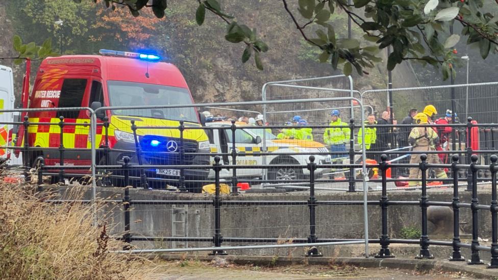 At least a dozen emergency service workers on the river bank in Sunderland, with a water rescue unit van in the foreground. There is temporary fencing around the area. Many of the emergency staff are wearing yellow high-viz jackets and helmets.