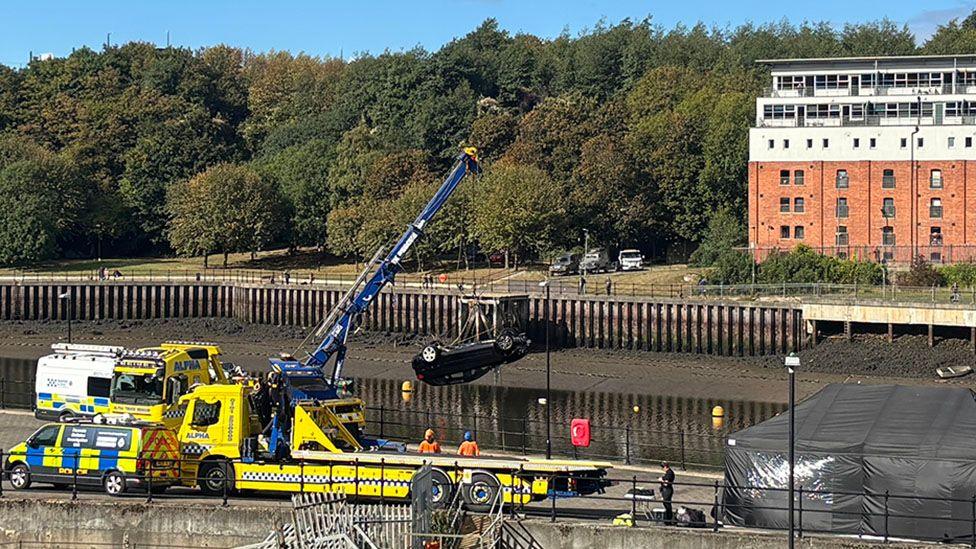 The River Wear in Sunderland with low water and brown, muddy banks, has a collection of emergency vehicles on the nearer quayside. There are marked police vans and a large, yellow recovery vehicle with a blue crane arm. Hanging from the crane arm is an upside down black Audi A3.
