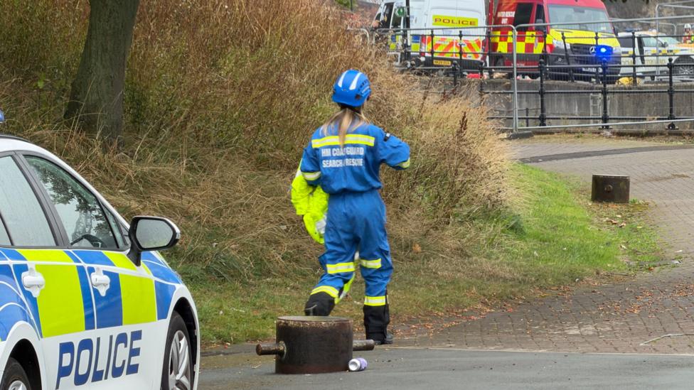 A woman in a blue HM Coastguard Search and Rescue suit walks away from a police car and towards the river bank, where other emergency vehicles are parked. The woman has blond hair in a ponytail and wears a blue helmet.