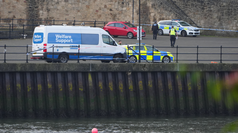 Around three emergency service workers walk on the river bank in Sunderland. There is temporary fencing around the area. Many of the emergency staff are wearing yellow high-viz jackets. There is a white van with 'welfare support' written on the side and two other police cars parked on the quayside.