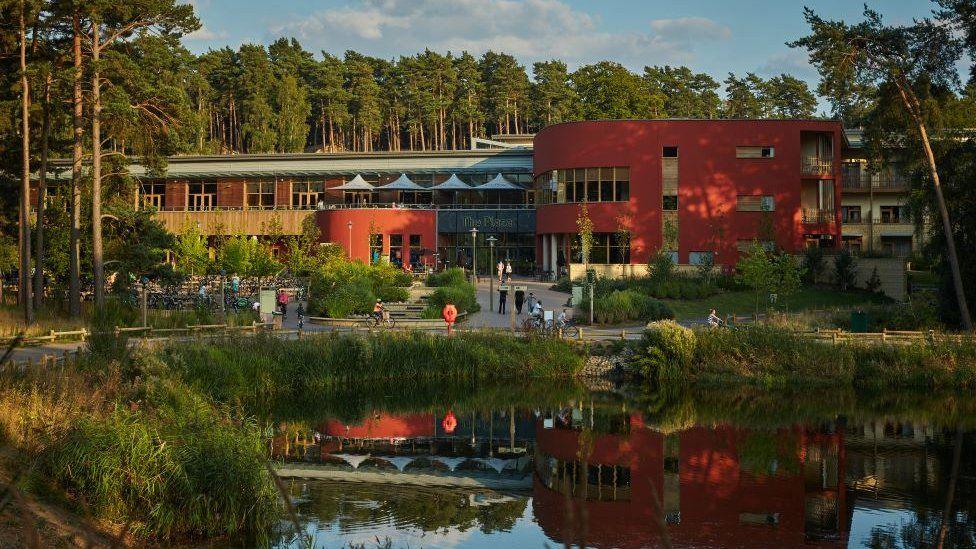 A lake edged by trees in front of a red-and-brown brick building. A reflection of the building is visible in the lake.