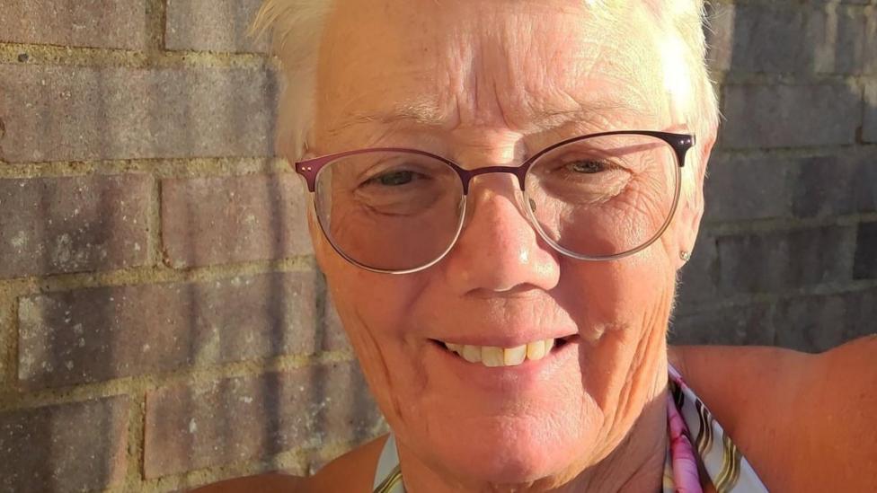 Penny Bulpitt: the face of a woman with short blonde hair and wearing brown-framed glasses. She is in front of brick wall and is smiling.