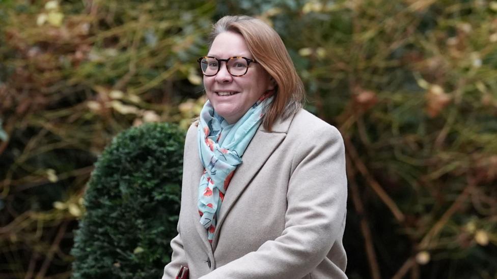A woman with dark hair, she is wearing glasses and a beige coat with a blue scarf