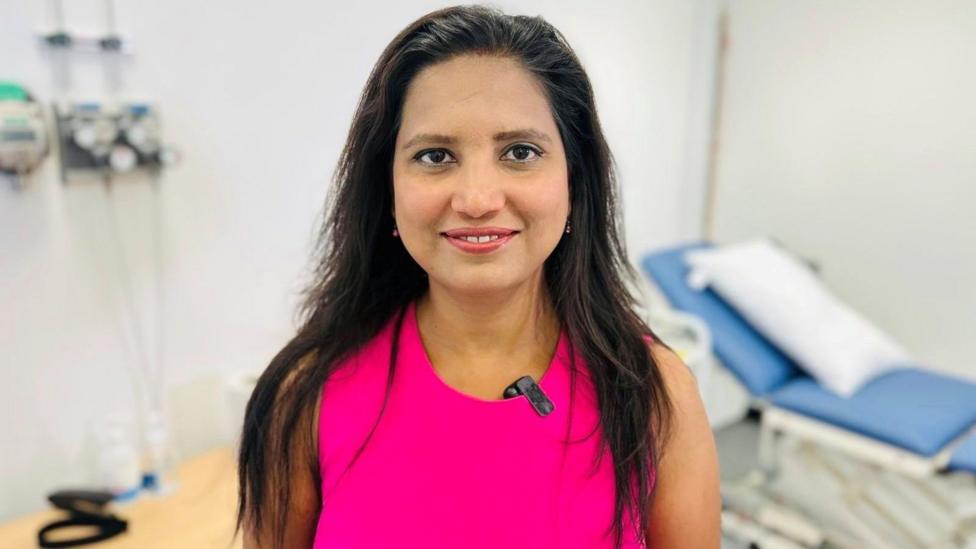 Dr Lucky Saraswat stands in a hospital setting with a bed in the background, she is smiling at the camera, and is wearing bright pink top.