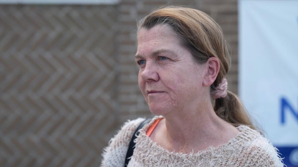 A white woman with brown hair looking off screen, she has her hair in a ponytail and is wearing a light brown jumper.