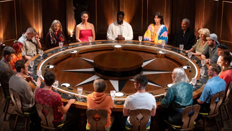 Contestants in series four of The Traitors sit around a large round table in the show's Round Table Room. The table is made of brown wood and has a black and white star-like decoration in its middle.