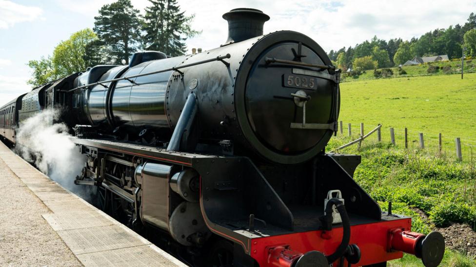 A black steam locomotive at a railway platform. Steam is rising from near its wheels. On the other side of the platform is a large grassy field.