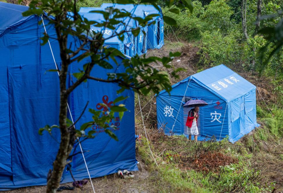 Photo: theukpulse.co.uk Blue disaster-relief tents on a muddy, forested hillside. A person holding an umbrella stands outside one of the tents