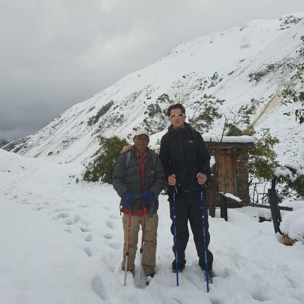 Jake Stacy and his Nepalese guide stand on a snow-covered mountain trail holding trekking poles, with steep white slopes rising behind them. A small stone hut partly covered in snow sits to one side of the path, while low clouds and an overcast sky surround the snowy landscape