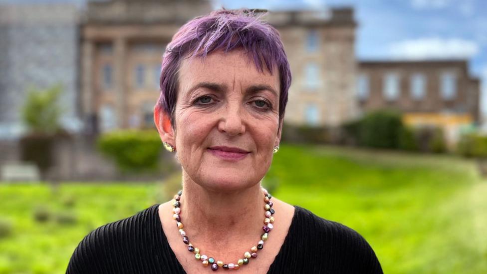 A woman with dyed purple hair in a black top and a necklace looks directly at the camera