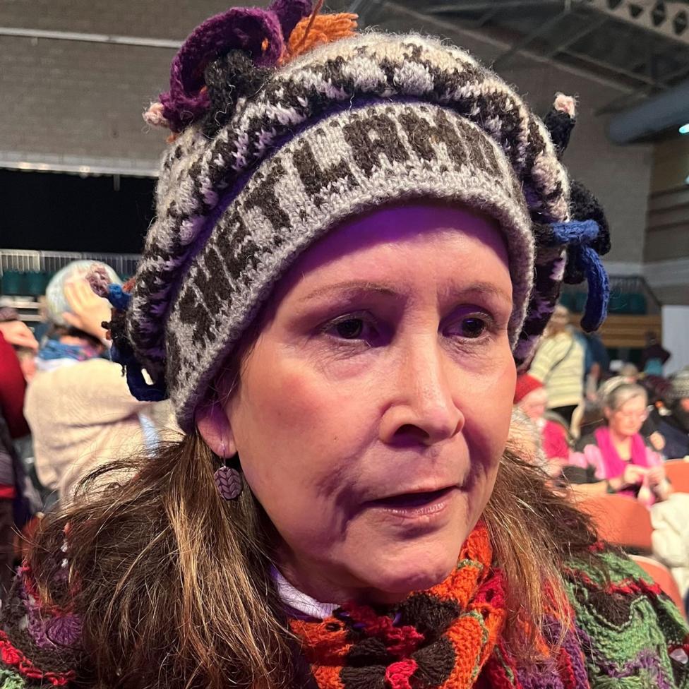 Nancy Woodhouse is wearing her hat over her long dark hair. The hat is woollen and grey in colour. The word "Shetland" is emblazoned along the head band. Nancy is standing in a large hall and there are people behind her.