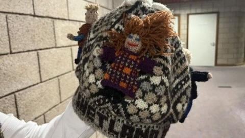 The picture has been taken indoors. Nancy is holding up the woolly hat which has ginger haired character stitched to a grey, pattern background.