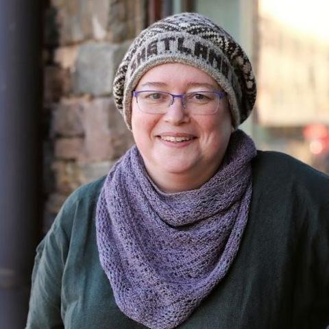 Helen is pictured outdoors. She is wearing a Birsie Beanny, lilac woollen scarf and a dark green top. She has glasses and is smiling.