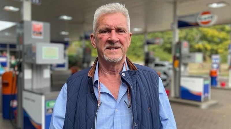 Ian Govier standing in the forecourt of Littlewick Green Motor Company in Maidenhead. He has short grey hair and a grey goatee beard. He is wearing a gillette over a blue shirt.