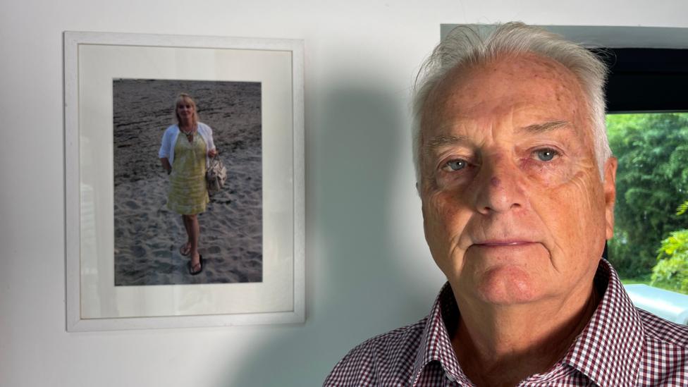 A man with grey hair wearing a pink checked shirt standing in front of a photo of a woman in a yellow dress who is wearing a white cardigan