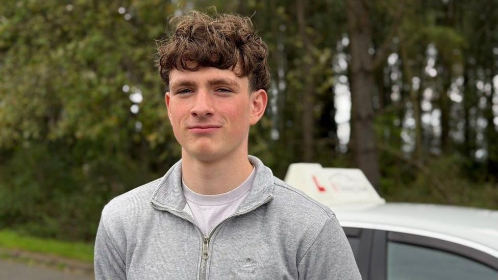 A young man with curly brown hair smiles with his mouth closed at the camera. He has a grey hoody on and a white top underenath