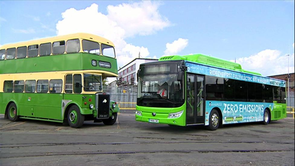 A green vintage double decker alongside a new electric bus parked on tarmac.