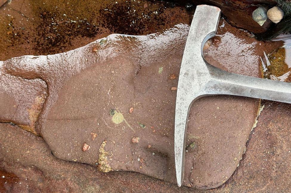 A close-up of red sandstone with different coloured markings in the rock at Clachtoll. Lying on top of the sandstone is a metal rock hammer.