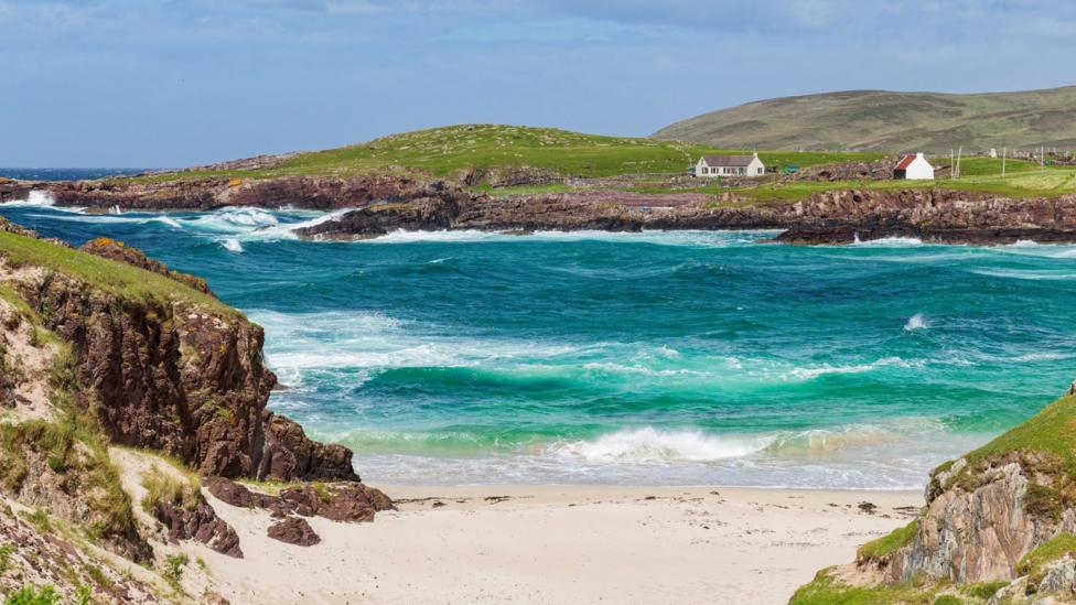 A beautiful landscape of rugged coastline and a patch of white sand. The sea is a bottle green colour and waves are breaking on the rocky shore.
