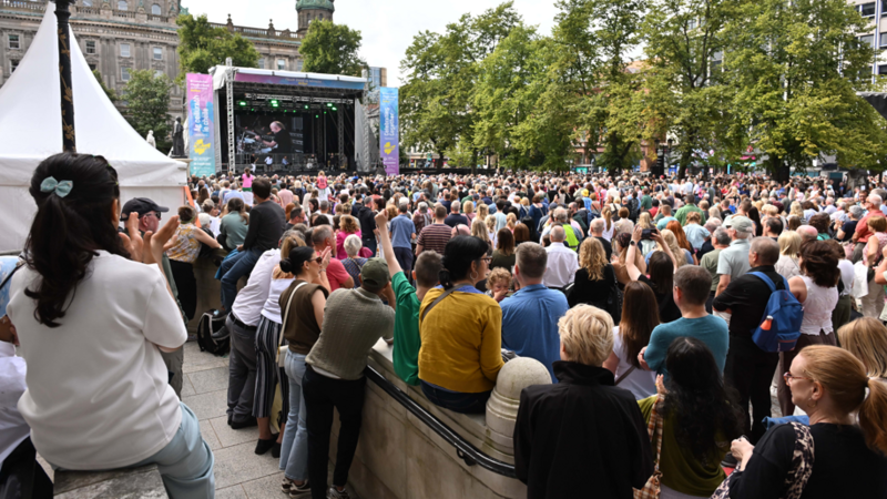A large crowd of people watch a performer on a stage outside Belfast City Hall. A screen above the stage shows the performer. Trees are at the front of the grounds. Tall, grey stone buildings are in the background.