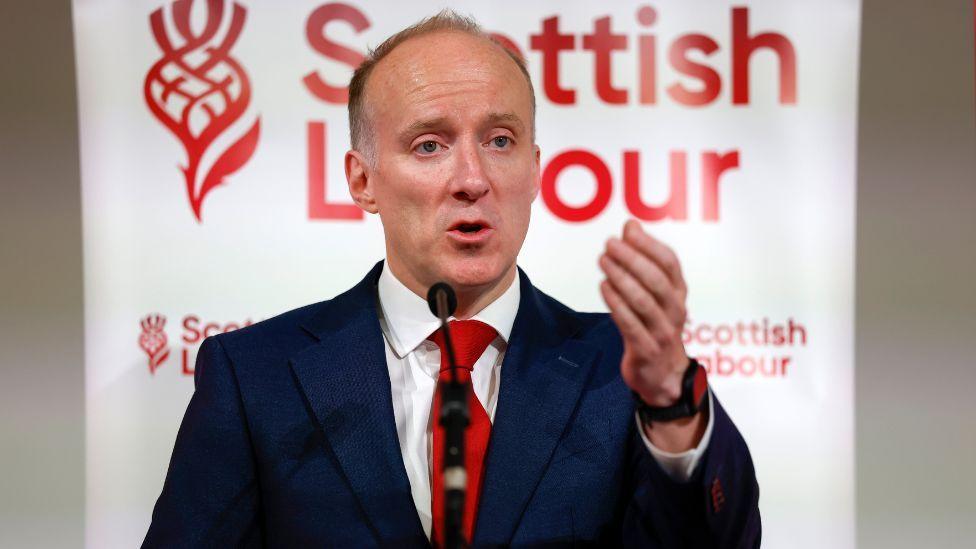 Michael Marra, who has thinning ginger hair, speaks into a microphone in front of Scottish Labour branding. He is wearing a business suit