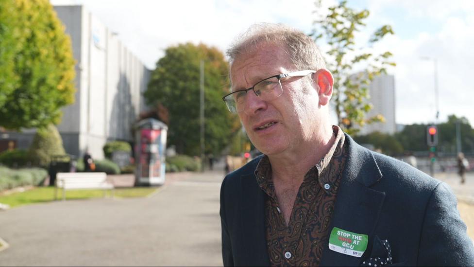 Person wearing glasses and a "Stop the Cuts at GCU" badge stands outside Glasgow Caledonian University campus during daylight, with university buildings and trees visible in the background.
