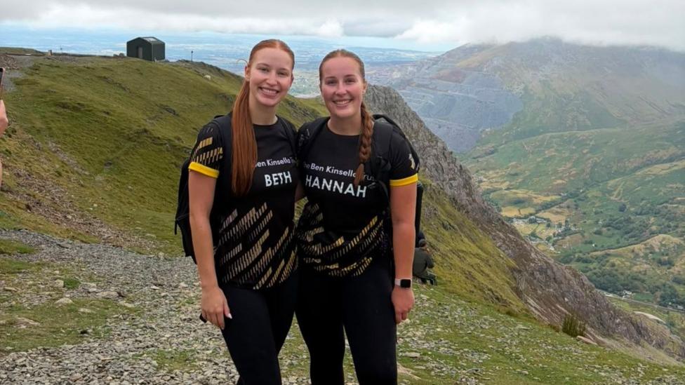 Two people wearing matching black charity challenge T-shirts and hiking boots stand on a rocky mountain path, posing for a photo. The landscape behind them shows steep green and rocky slopes, a valley below, and distant mountains partially covered by low cloud and mist under an overcast sky. A small building can be seen on the hillside in the background.