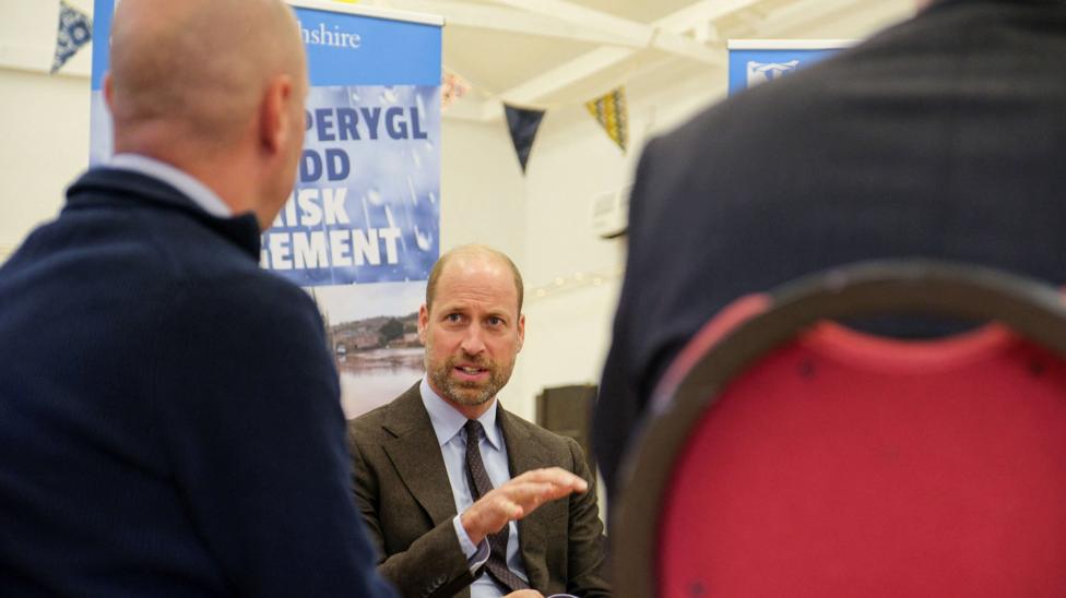 Prince William in a suit sat on a chair. Can see the backs of two men facing him sat on red chairs in what looks like a hall.