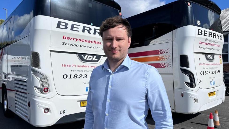 James Berry is wearing a blue shirt and standing in front of two white coaches with the name 'Berry's' on the back