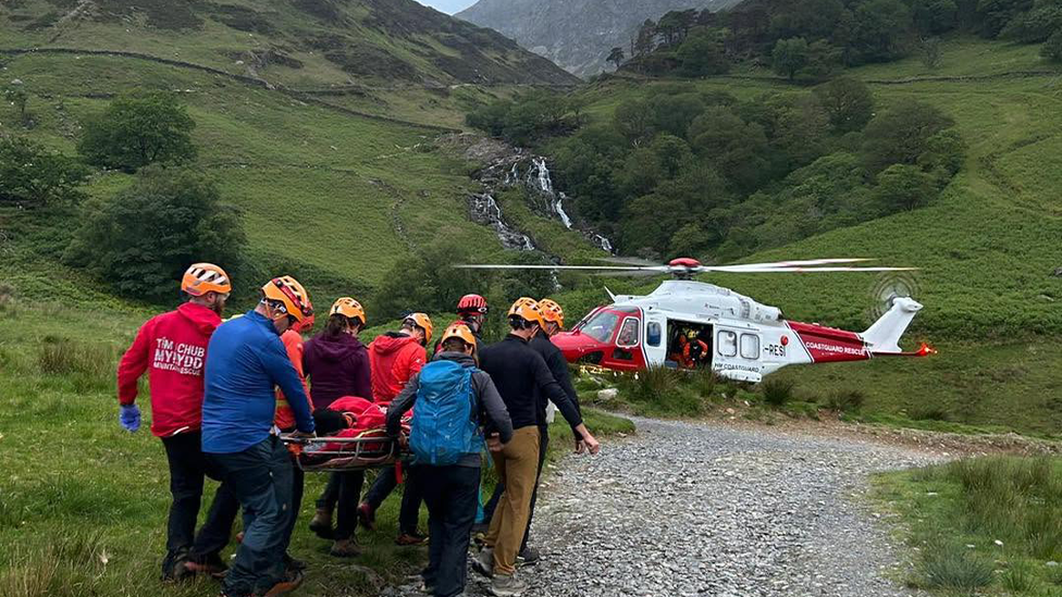 About ten mountain rescue volunteers in yellow safety helmets carry a person on a stretcher down a gravel path towards a Coastguard Rescue helicopter in a remote valley. A waterfall and steep green hills can be seen in the background.