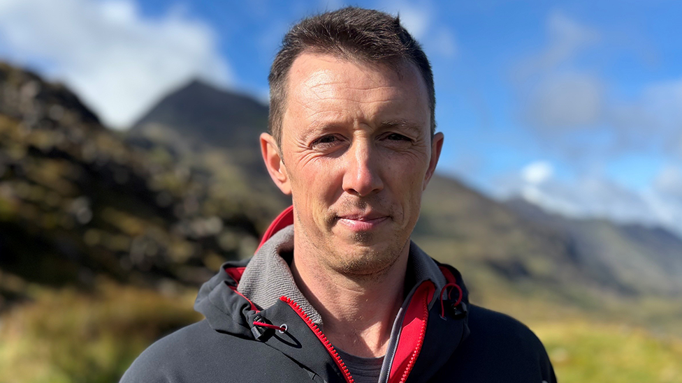 Tom Carrick is wearing an outdoor jacket stands in front of a mountainous landscape under a blue sky. Rocky slopes and grassy ground are visible in the far distance behind him.