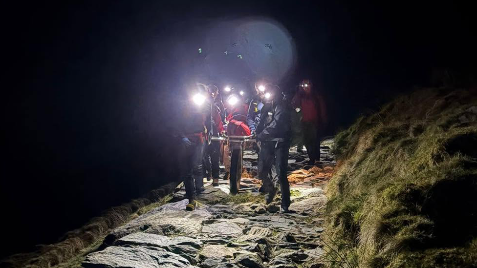 Mountain rescue volunteers wearing head torches carry a person on a stretcher along a rocky path at night. The surrounding hillside is dark, with the route lit only by the volunteers’ lights which are causing a halo effect .