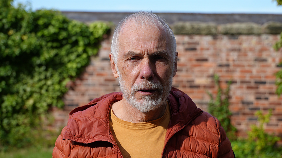 Caradoc Jones with short grey hair and a white beard stands outdoors wearing an orange jacket over a yellow T-shirt. A brick wall and green foliage are visible in the near background with blue sky in the distant background