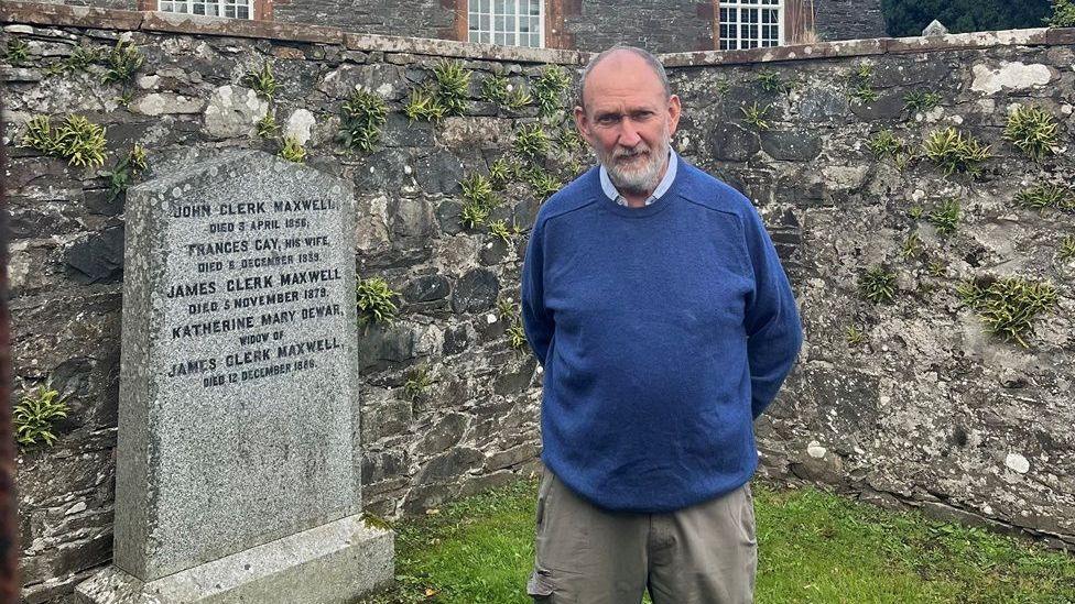 A man in a blue jumper with receding grey hair and a beard stands in front of the gravestone of James Clerk Maxwell