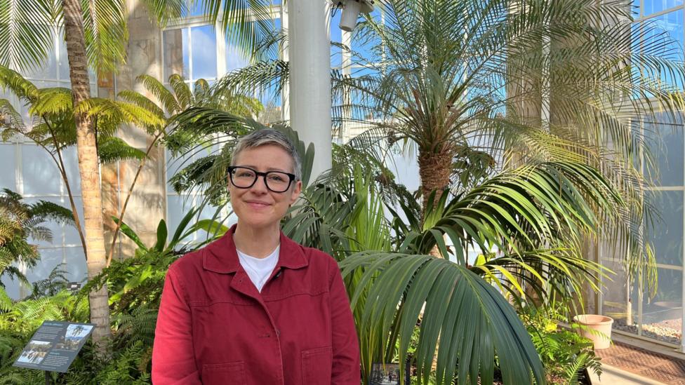A smiling, short-haired woman with black rimmed glasses and a red jacket smiles standing in front of a palm tree, glass windows behind her.