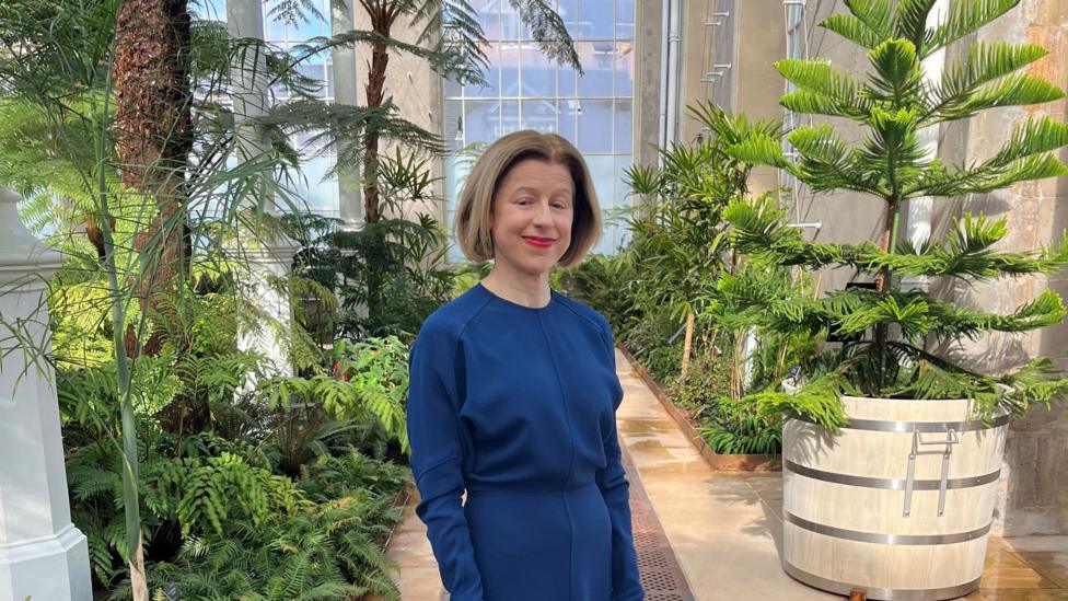 A smartly dressed woman in a blue tailored dress, and neat brown hair in a bob stands in a path that leads back into the palm house.
