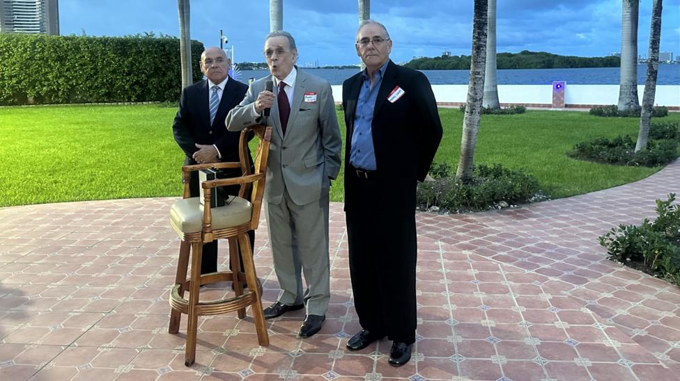 Juan Omar Sixto speaks into a microphone, stood between two of his colleagues. They are standing in front of a stool on which is a speaker