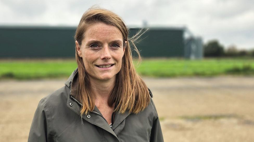 A woman standing outdoors in a rural setting, wearing a dark weatherproof jacket. A gravel surface, green grass and large agricultural buildings appear softly blurred in the background beneath an overcast sky.