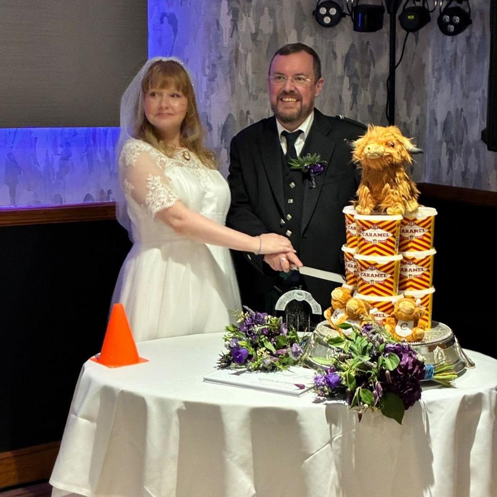 A bride and groom cut their wedding cake which sits on a table and is made of tubs of caramel logs, with a highland cow on top.