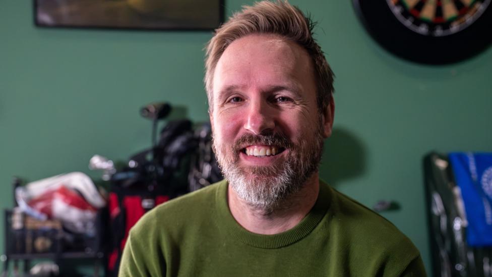 A close-up shows a man seated indoors, wearing a green sweater, with golf clubs visible in the background. The room has green walls and sporting touches, including a dartboard, creating a relaxed, personal setting focused on golf and leisure activities.