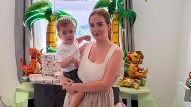 Sarah Durgan holds her son Noah in her arms on her left hip. She wears a pink dress and smiles at the camera. She has brown hair and blue eyes. Noah wears a white t-shirt and shorts and has curly hair. Dinosaur balloons and palm trees can be seen on the table behind them.