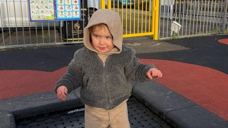 Photograph of Noah in a grey teddy fleece and brown hooded jacket and trousers. He stands on a trampoline in a playground and smiles. He has blue eyes and the sun shines on his face.
