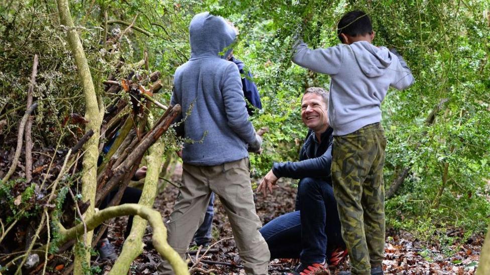 Steve Backshall crouches among trees and undergrowth in a wooded area while children stand nearby. Fallen leaves cover the ground and a den-like structure built from branches and sticks is visible to one side. The children are taking part in an outdoor activity.