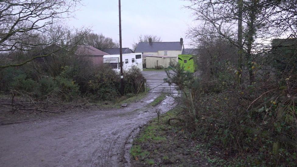 The entrance gate to a farm can be seen with a green horse trailer on the right.