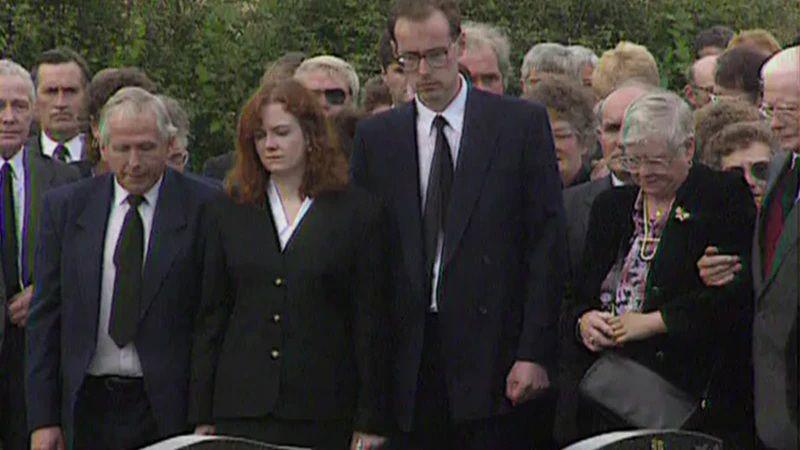 People attend a funeral in formal attire. Centrally a woman with ginger hair and a tall man with brown hair and glasses look down at the coffin with forlorn expressions on their faces.