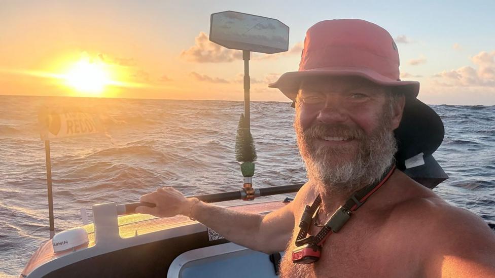 Dave Bell smiles for a selfie in his boat as the sun sets on the ocean horizon. He is wearing a red sun hat and is bare chested. He has a full, grey beard and is wearing an electronic device around his neck.