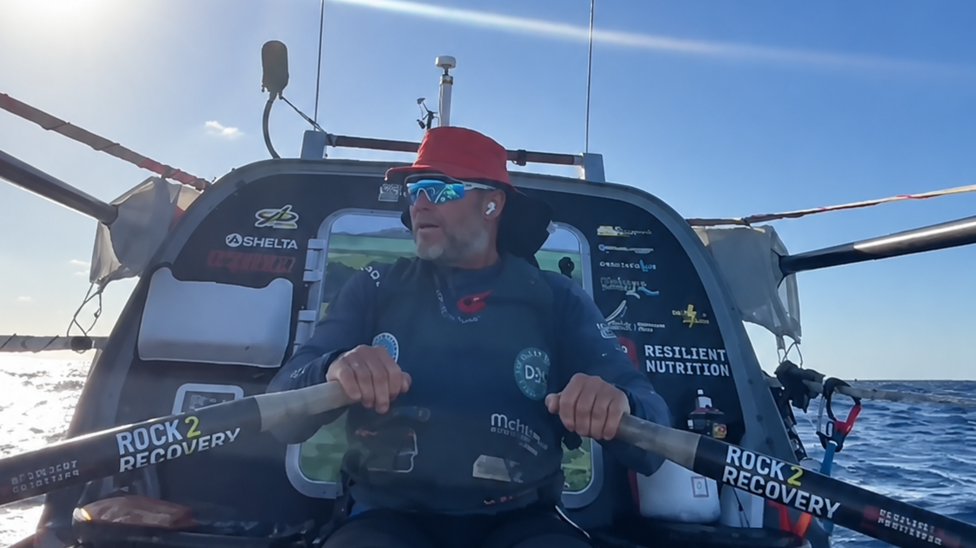 Dave Bell holding the oars of his ocean rowing boat. He is wearing a red sun hat, a long-sleeve top bearing sponsor logos and reflective sports sunglasses.