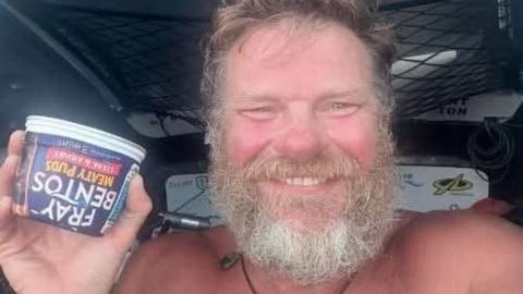 Bell has a wide smile as he holds up a Fray Bentos tinned pie. He has a grey beard and is not wearing a shirt.