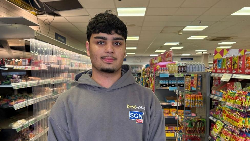 Hakim has black curly hair and wears a grey hoody with best-one and SGN retail embroidered on it. He stands in the shop with the aisles behind him. 